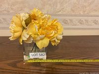 Yellow artificial flower arrangement in clear glass rectangular container placed on wooden surface with measuring tape showing approximate length.