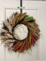 Front view of wreath showing beige, brown, and red artificial plant clusters arranged circularly with green leaves and thin brown stalks on a white door, labeled Lot 30, showing size tag.
