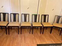Front view showing six wooden dining chairs with beige upholstered seats lined up on wood flooring in front of a closet space.
