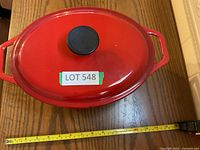 Top view of red enamel cast iron cookware with lid, showing black knob handle and LOT 548 label with measuring tape placed below showing approx 14 inches.