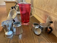 Photo showing two aluminum stovetop espresso makers and one red glass pitcher on a wooden surface. Smaller espresso maker is assembled, larger one disassembled, pitcher has textured red glass with handle and lid.