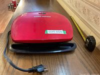 Red George Foreman grill on wood surface with tape label 'LOT 554' and measuring tape beside it showing approximate size.