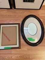 Two picture frames side by side on a wooden floor - one oval antique frame with curved glass and matting, and one square wooden frame without glass.