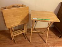 Four wooden TV trays with folding legs and a wooden stand against a wall on a wood floor.