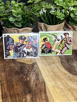 Two 1966 Batman Topps trading cards displayed side by side with leafy plants in the background.