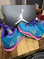 Pair of Jordan Retro 5 baby shoes in size 4C displayed on a wooden surface in front of black box with white Jumpman logo. Blue, pink, and purple color scheme visible.