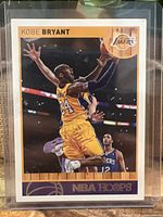Front of Kobe Bryant 2013 NBA Hoops card in protective sleeve showing Kobe in mid-air with Lakers uniform number 24 during a game