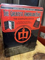 Close-up photo of the front of the sealed tin showing The Greatest American Hero Limited Collectors Edition complete series with images of cape, iron-on, and manual.
