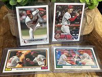 Four baseball cards displayed in plastic sleeves on a table with plants in the background. The cards feature Shohei Ohtani, Mike Trout, and Juan Soto.