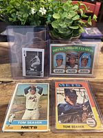 Four vintage baseball cards displayed on wood table with greenery in background. Cards include 8 of Diamonds black and white card, 1971 Strikeout Leaders card, 1975 and 1976 Topps cards of Tom Seaver.