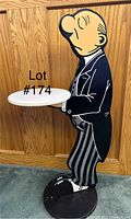 Photo of wooden butler figure designed as an end table holding a round white tray, standing against a wood-paneled wall.