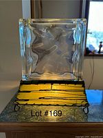 Unique glass block lamp with clear textured glass block and decorative golden metal base, photographed on a table in natural lighting.