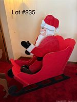 Side view of the wooden Santa Claus figure seated in a large red sleigh with black runners, showing details of the painted face, hat, and gloves.