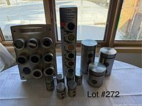 Photo showing a flat stainless steel spice rack holding eight round spice jars with various spice contents, a vertical rotating spice rack with ten small spice jars, and four stainless steel canisters with lids arranged on a white tablecloth in front of windows.