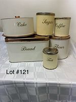 Six vintage kitchen metal canisters labeled Cake, Sugar, Coffee, Bread, Flour, and Tea. All have lids, some lids appear copper-colored. The canisters show light wear and vintage style script labeling.