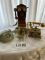 Five vintage telephones arranged on a tablecloth. Includes a wooden Thomas museum series wall phone with brass bells, two rotary dial phones, one with cream color and one beige, an ornate brass phone with decorative elements, and a light green rotary dial phone.