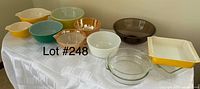 Photo showing a collection of ten vintage Pyrex bowls and casserole dishes of various colors and sizes arranged on a white tablecloth.