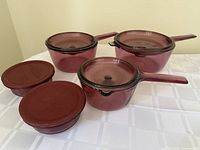 Photo of three purple glass saucepans with handles and lids, plus two smaller round containers with maroon plastic lids, all placed on a white tablecloth.