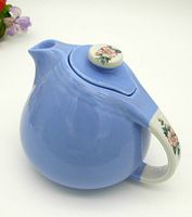 Blue ceramic teapot with Rose Parade floral decoration on handle and lid knob, side view showing spout and rounded body.