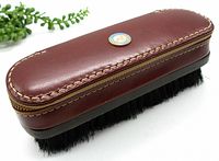 Closed leather travel valet with rounded shape, brown leather, stitching along edges, and black brush bristles underneath.