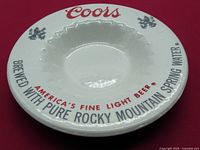 Top view of the white ceramic dish showing red Coors logo, black and red printed text around rim, and pine tree graphics.
