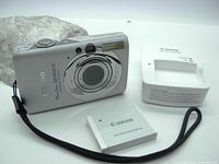 Photo showing the Canon PowerShot SD1200IS camera with battery pack NB-6L and battery charger, all placed on a white surface.