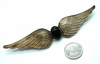 Large gold-tone metal wings brooch with detailed feather engraving and central green oval cabochon stone, next to a coin for scale.