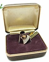 Tie tack displayed in its original hinged case with a brown leatherette exterior, gold-toned satin interior lid, and purple velvet base.