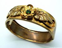 Close-up angled view showing raised flower with green stone center and surrounding leaf design on gold tone bangle bracelet.