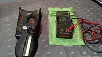 Fluke digital multimeter with red and black test leads on green anti-static bag, next to vintage clamp meter in leather case on fabric surface.