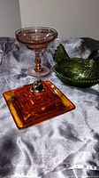 Green depression glass hen covered dish with detailed feathers and beaded edge, pink depression glass compote dish with decorative stem, and amber Viking pyramid ashtray on a silver-gray fabric background.