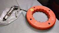 Orange plastic reel containing flat steel fish tape, 200 feet long and 1/8 inch wide, placed next to the grease gun on a surface.