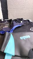 Photo showing four wetsuits arranged with Omba wetsuit in foreground with blue panels and zipper visible; additional wetsuits behind including black and blue colors.