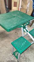 Full view of the green plastic picnic table with attached seating in a folded or semi-folded state in a garage or storage area.