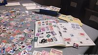Wide view of collector's book open with multiple USA stamps affixed in slots and many loose stamps scattered around on a table.