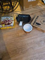 Photo showing boxed La Forme Deep Dish Swiss Rosette 11-inch bakery pan with recipe booklet, small ceramic bowl with copper holder, black rectangular pan and stoneware pie plate on floor.