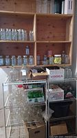 Overall view of wooden shelving with multiple glass jars, metal lids, and boxed items including large glass carboys on bottom shelf.