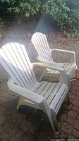 Two white plastic Adirondack style outdoor chairs placed on brick surface under shade, showing weathering and dirt marks.