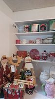 Photo showing a white cabinet interior with multiple Christmas-themed ceramic items, including mugs, plates, two figurines (reindeer and snowman with sign), and a miniature wagon with trees on the lower shelf.