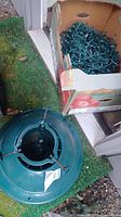 Photo showing green plastic Christmas tree stand and partially open box with Christmas lights inside.