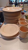Stack of beige ceramic plates in two sizes along with five matching beige ceramic mugs placed on a kitchen counter.