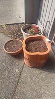 Three flower pots grouped outdoors on pavement: a large scalloped square terracotta pot decorated with floral designs, a smaller round terracotta pot, and a larger beige plastic pot with red flowers planted.