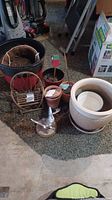 Photo of assorted flower pots, including terracotta, ceramic and plastic pots, small wicker chair garden decor, and a metal bird figurine on stand all arranged on carpet.