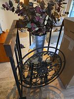 Large welded metal circular wall art with scroll and floral design, black metal stool, and galvanized metal vase with artificial eucalyptus stems shown from front angle.