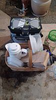 Photo showing picnic basket filled with white ceramic coffee carafe, plasticware, and containers, with cooler bag and plastic jug behind