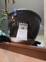 Glossy black ceramic plant pot shown on a wooden ledge, with attached matching water tray underneath. Pot contains soil and remains of a house plant with some dead stems and leaves visible.