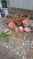 Photo of outdoor area showing 12 terracotta pots of various sizes, some containing soil inside, placed on a gravel and mossy patch near a building wall.