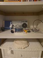 Stereo cassette recorder with speaker, photo book, tape dispenser, crayons, electronic game, storage tin, toy truck, and metal airplane bank on shelf with wicker basket below.