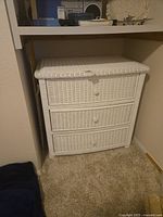 Photo showing the front and side angle of the white wicker three drawer dresser fitted into an alcove space. Drawers have small round wicker knobs and appear to slide smoothly on metal tracks.