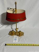 Front view of the antique French bouillotte lamp showing the red metal shade with gold painted decoration and the brass base with two candle sockets; measuring tape placed for scale.